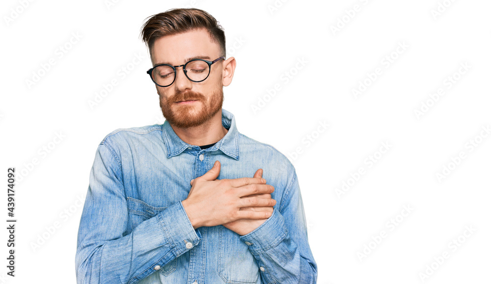 Young redhead man wearing casual denim shirt smiling with hands on chest with closed eyes and grateful gesture on face. health concept.