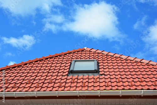 Roofing construction. A red metal roof with a attic skylight window installed, a plastic rain gutter, a fascia board and a soffit against the blue sky.