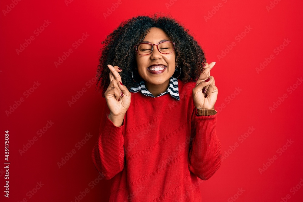 Beautiful african american woman with afro hair wearing sweater and glasses gesturing finger crossed smiling with hope and eyes closed. luck and superstitious concept. Beautiful african american woman with afro hair wearing sweater and glasses gesturing finger crossed smiling with hope and eyes closed. luck and superstitious concept.