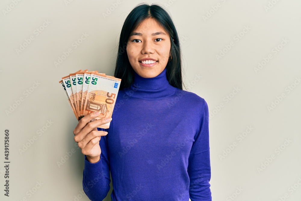 Young asian woman holding bunch of 50 euro banknotes looking positive and happy standing and smiling with a confident smile showing teeth Young asian woman holding bunch of 50 euro banknotes looking positive and happy standing and smiling with a confident smile showing teeth