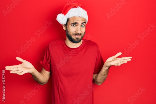 Young hispanic man wearing christmas hat clueless and confused expression with arms and hands raised. doubt concept.