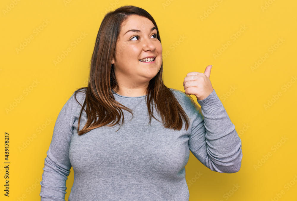 Beautiful brunette plus size woman wearing casual clothes smiling with happy face looking and pointing to the side with thumb up. Beautiful brunette plus size woman wearing casual clothes smiling with happy face looking and pointing to the side with thumb up.