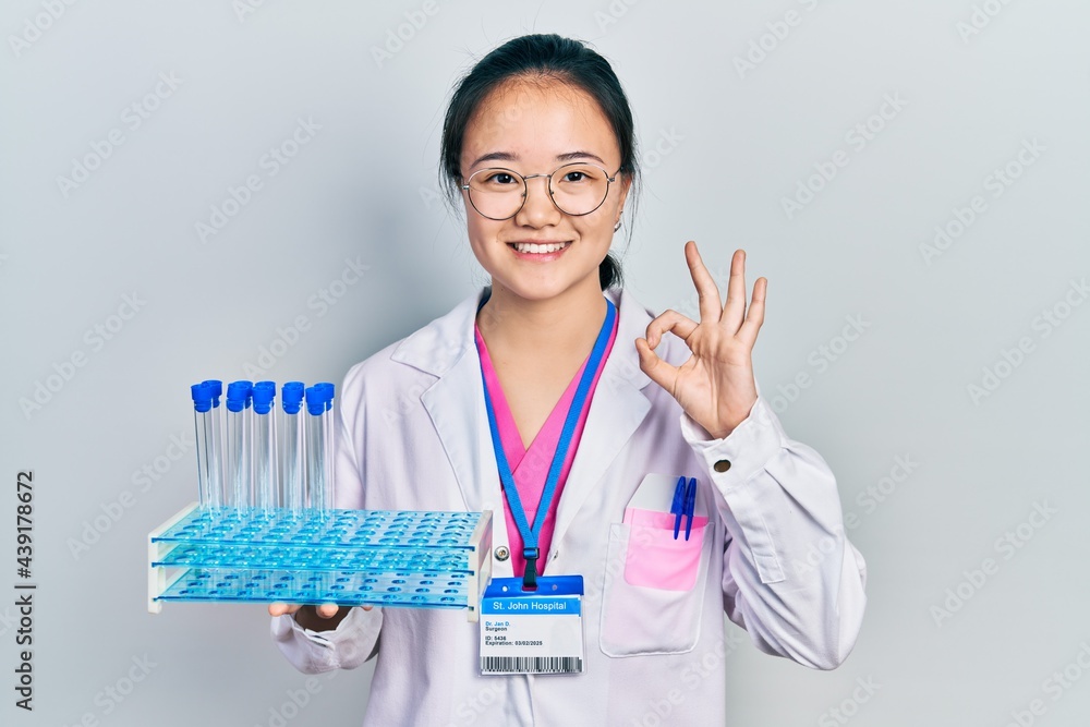 Young chinese girl wearing scientist uniform holding test tube doing ok sign with fingers, smiling friendly gesturing excellent symbol Young chinese girl wearing scientist uniform holding test tube doing ok sign with fingers, smiling friendly gesturing excellent symbol