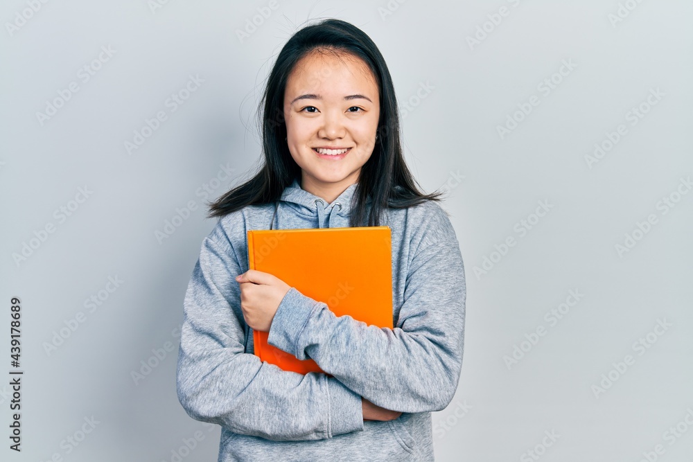 Young chinese girl holding book smiling with a happy and cool smile on face. showing teeth. Young chinese girl holding book smiling with a happy and cool smile on face. showing teeth.