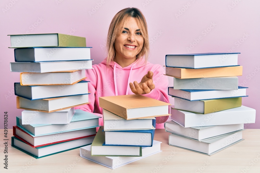 Young caucasian woman sitting on the table with books smiling cheerful offering palm hand giving assistance and acceptance. Young caucasian woman sitting on the table with books smiling cheerful offering palm hand giving assistance and acceptance.