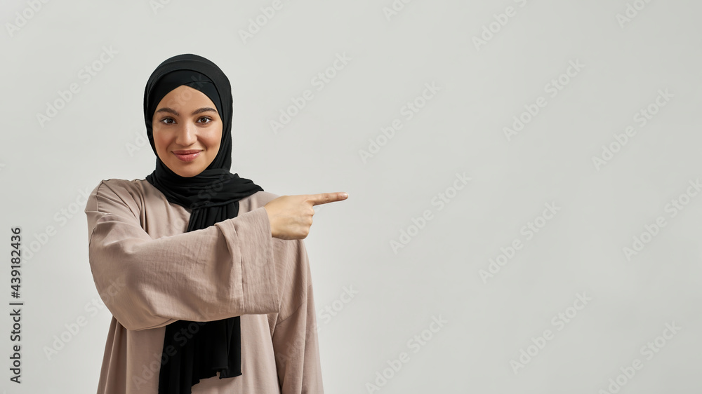 Smiling young arabic woman in hijab pointing to side - obrazy, fototapety, plakaty 