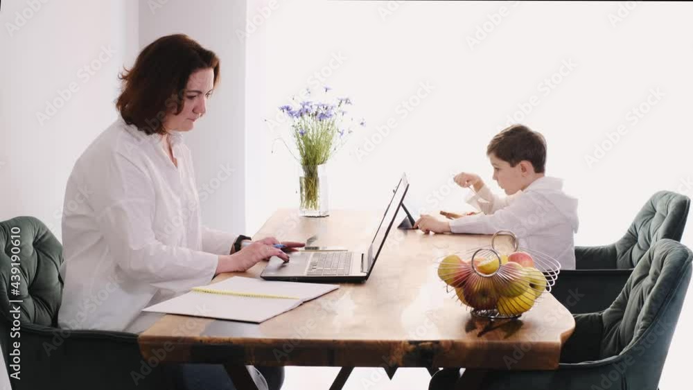 Middle age woman working at home on laptop while child is eating food on the same table with tablet. Working mother with son using gadgets on wooden table kitchen. Busy freelancer, distant employee Middle age woman working at home on laptop while child is eating food on the same table with tablet. Working mother with son using gadgets on wooden table kitchen. Busy freelancer, distant employee
