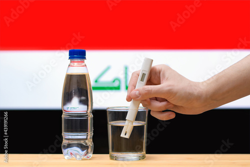 A hand with a water tester makes a measurement in a glass of clear water against the background of the flag of Iraq. Test and assessment of drinking water supplies in Iraq.