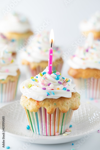 A single confetti cupcake with a lit candle, on a plate with several cupcakes in soft focus in behind.