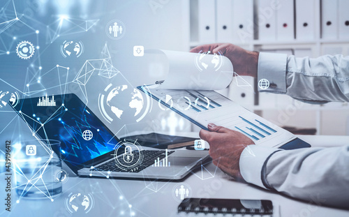 Businessman sitting at workplace in front of laptop and checking report with graphs. Office in the background. Digital interface in the foreground. Concept of data internet security in networking