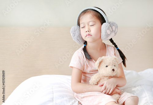Fototapeta Naklejka Na Ścianę i Meble -  Adorable little Asian child girl wearing winter earmuffs and hugging teddy bear while sitting on the bed at home.