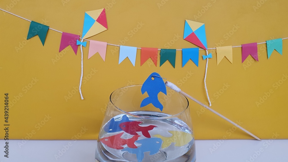 Kites, flags, colorful fish in the jar with water, fishing rod, on the table. Yellow background. Tradition to brighten up the Brazilian June party is the play and fun of toy fishing. Space for text. - obrazy, fototapety, plakaty 