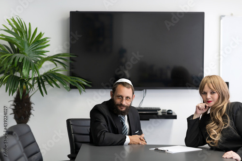 Two Israeli business partners is working, sitting at a desk in the office. Jewish man in a yarmulke and glasses sitting with woman with blond hair. Jew man in shirt, suit and national hat Kippah