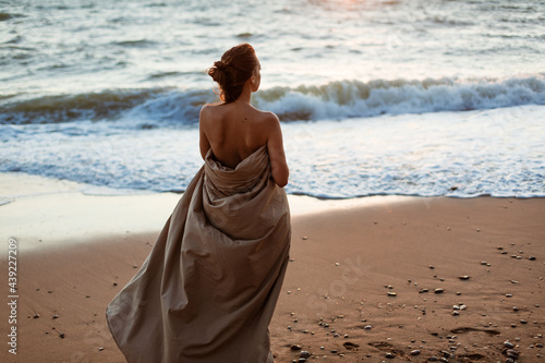 Beautiful young woman wrapped in a blanket. The girl on the seashore in the evening stands. Lonely looking into the distance on the beach at sunset. Summer evening by the water