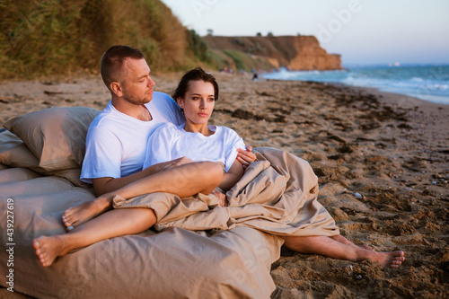 Young couple wrapped in a blanket outdoors. Happy couple in love lying and rotating in bed by the sea at sunset. Happy caucasian couple of husband and wife wrapped in a blanket outdoors
