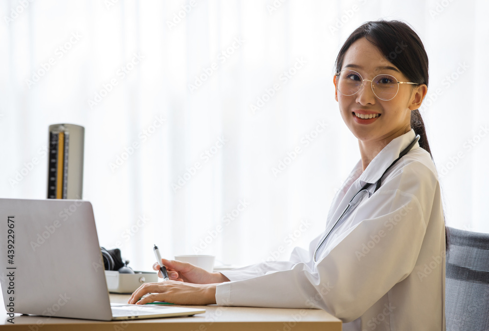 Young asian optometrist who wear cute eyeglasses in white uniform sit in the office clinic in hand holding pen and touch laptop. Seems busy but still have beautiful smile to camera man. Young asian optometrist who wear cute eyeglasses in white uniform sit in the office clinic in hand holding pen and touch laptop. Seems busy but still have beautiful smile to camera man.