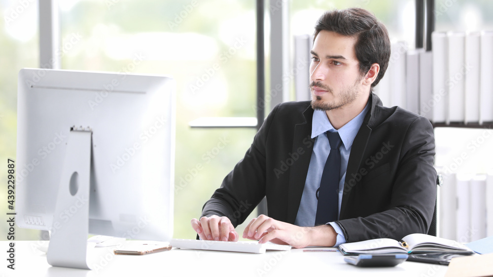 Good looking young adult businessman in suit sitting at desk and working with desktop computer in modern office Good looking young adult businessman in suit sitting at desk and working with desktop computer in modern office
