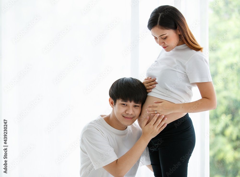 young lovely Asian husband and wife stay together in the living room. A pregnant woman standing and looking at her belly while a man sitting and listening to a baby in her belly with love copy space young lovely Asian husband and wife stay together in the living room. A pregnant woman standing and looking at her belly while a man sitting and listening to a baby in her belly with love copy space
