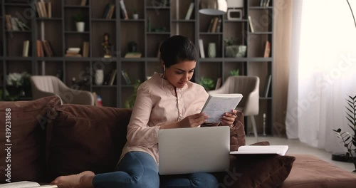 Indian ethnicity female student makes assignment while sit on sofa with laptop on laps, holding copybook writes notes listens audio course through wired earphones. E-learning process at home concept