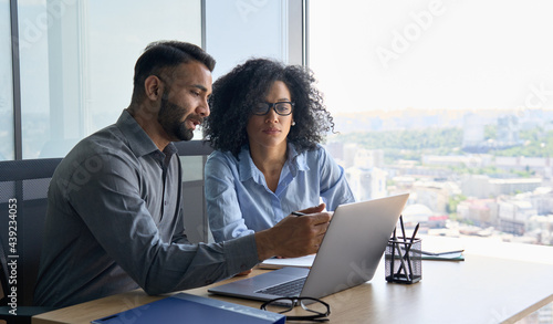 Indian male ceo executive manager mentor giving consultation on financial operations to female African American colleague intern using laptop sitting in modern office near panoramic window. photo