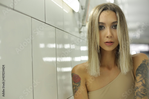 portrait of an interesting young woman in a beige swimsuit and with tattoos on the body which stands indoors with a white tile on the walls and looks directly into the camera. High quality photo