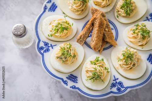 Kalte gefüllte russische Eier mit Frischkäse, Kresse und Kräuter farciert auf Blau Weiß Porzellan Eierplatte, Leinen Tuch und Marmor Hintergrund hell mit Salzstreuer und Toast Brot 