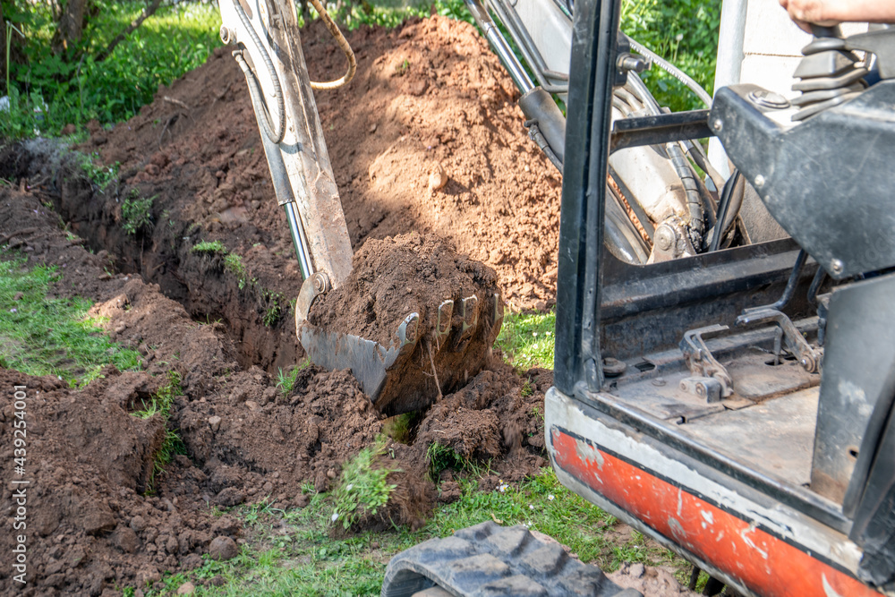 Fototapeta premium Mini excavator digs the ground. Trench for water pipes or communication lines