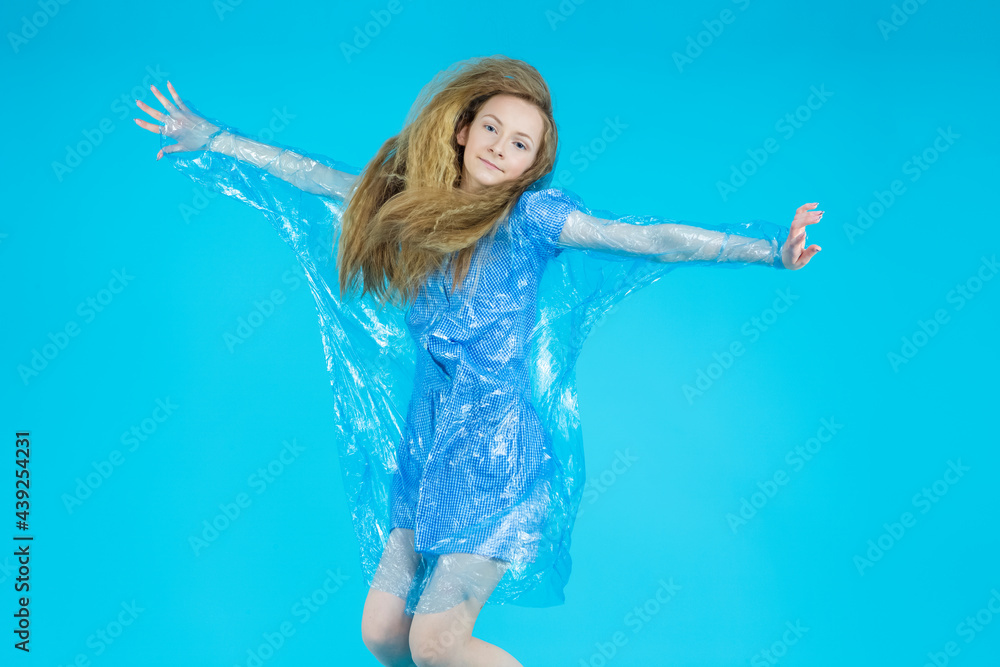 Teenager Lifestyle. Active Teenage Girl In Blue Dress, Transparent Raincoat and Rubber Boots Moving in Jump Against Blue Background.