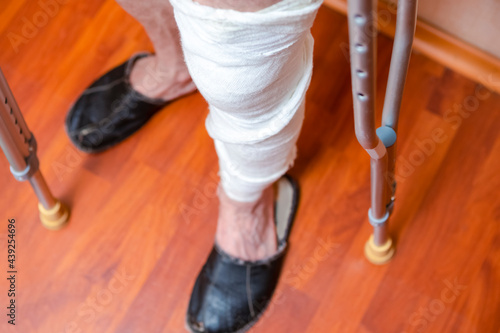 Medical Ideas. Disabled Man With Plaster And Bandage on His Leg Standing With Pair of Crutches Indoors.