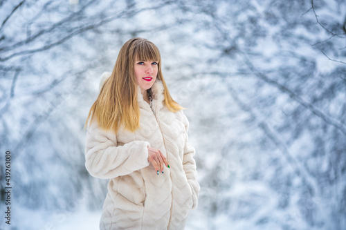 Beautiful smiling confident young white woman pretty face in a white fur coat looking at camera posing alone at snowy forest