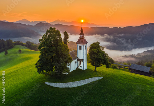 Skofja Loka, Slovenia - Aerial view of the beautiful hilltop church of Sveti Tomaz (Saint Thomas) with amazing golden foggy sunrise and the Julian Alps at background at summer time