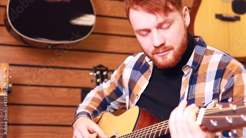 Irishman redhaired bearded handsome man in brown plaid casual shirt choosing a guitar in a music store