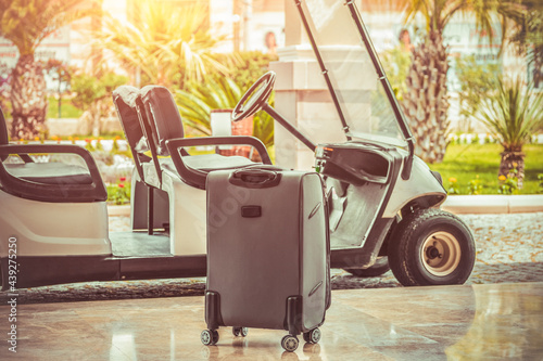 Fototapeta Naklejka Na Ścianę i Meble -  A suitcase standing near electrical hotel passenger shuttle bus in front of blurry palm trees. Summer holiday concept with golf car service and luggage.