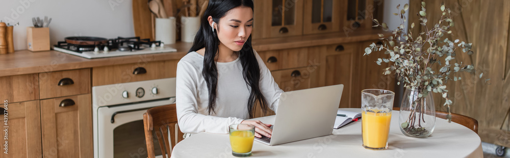 serious young asian woman in earphones sitting on table and using laptop near notebook with pen in kitchen, banner serious young asian woman in earphones sitting on table and using laptop near notebook with pen in kitchen, banner