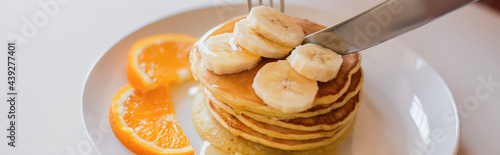 close up view of knife cutting pancakes with orange and banana slices on white plate, banner