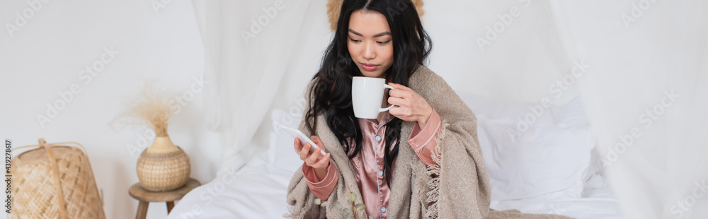 young asian woman in silk pajamas and blanket sitting on bed, drinking coffee and holding cellphone in bedroom, banner young asian woman in silk pajamas and blanket sitting on bed, drinking coffee and holding cellphone in bedroom, banner