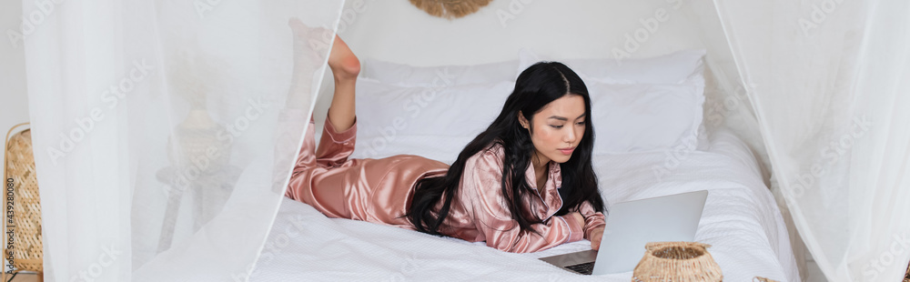 young asian woman in silk pajamas lying on bed with laptop in modern bedroom, banner young asian woman in silk pajamas lying on bed with laptop in modern bedroom, banner