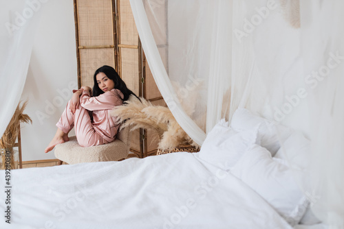 sad young asian woman sitting on chair in self hug pose and looking at camera in bedroom © LIGHTFIELD STUDIOS