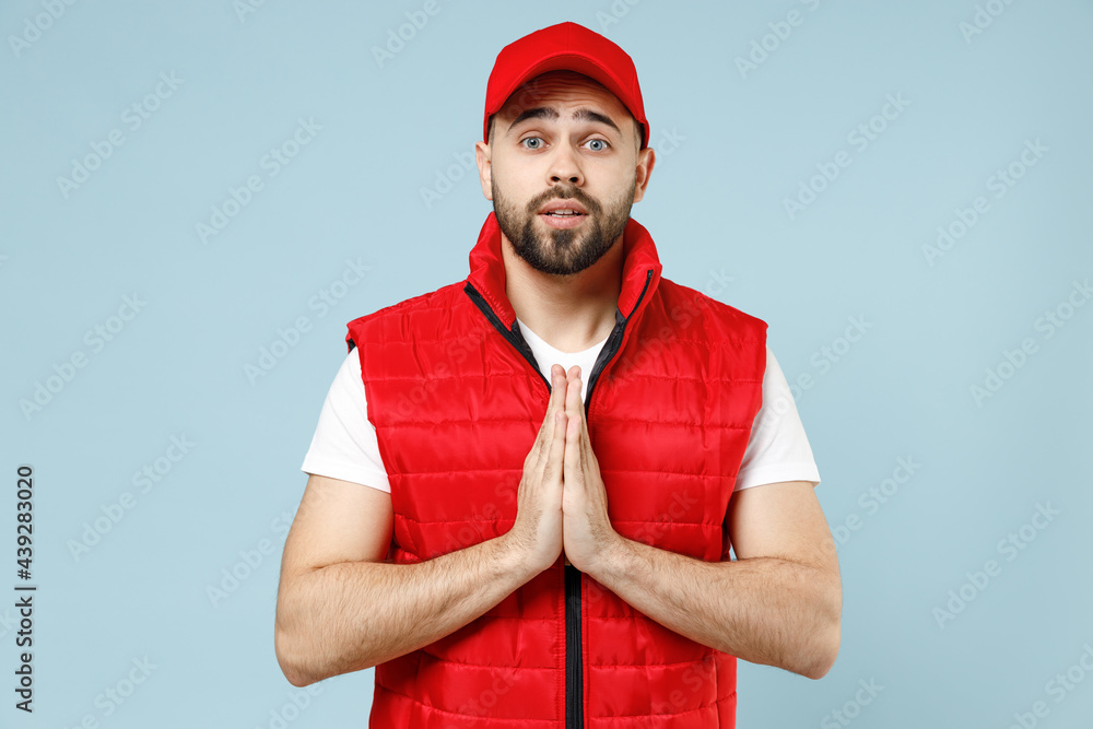 Professional delivery guy employee man in red cap white T-shirt vest uniform workwear work as dealer courier begging about something isolated on pastel blue color background studio. Service concept. Professional delivery guy employee man in red cap white T-shirt vest uniform workwear work as dealer courier begging about something isolated on pastel blue color background studio. Service concept.