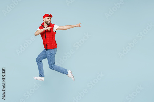 Full size body length side view delivery guy employee man in red cap white T-shirt vest uniform work dealer courier jump run go walk isolated on pastel blue color background studio. Service concept