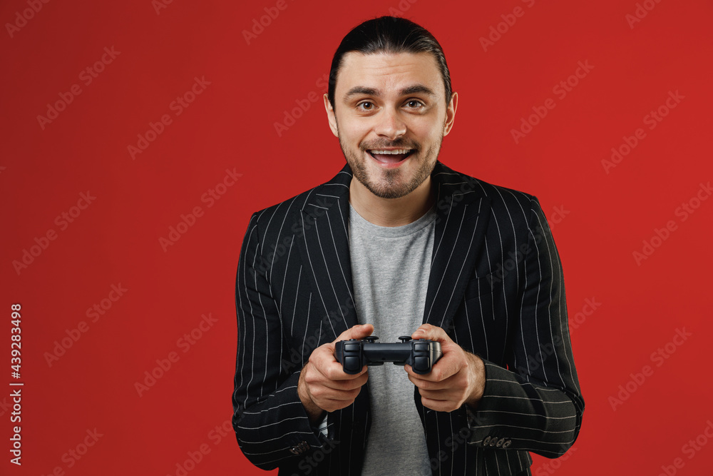 Young excited happy joyful smiling fun successful unshaven latin man 20s wearing black striped jacket grey shirt play pc game with joystick console isolated on red color background studio portrait Young excited happy joyful smiling fun successful unshaven latin man 20s wearing black striped jacket grey shirt play pc game with joystick console isolated on red color background studio portrait