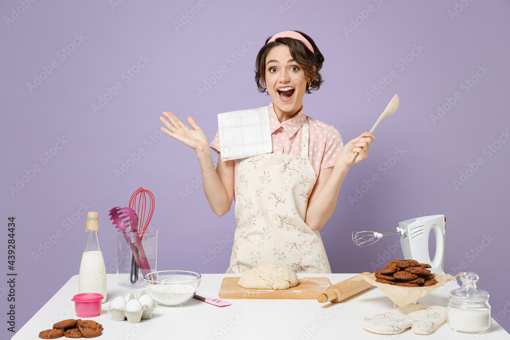 Young excited amazed housewife cook chef baker woman wear pink apron work at table kitchenware hold wooden spoon spread hands isolated on violet background studio Process cooking food pastry concept. Young excited amazed housewife cook chef baker woman wear pink apron work at table kitchenware hold wooden spoon spread hands isolated on violet background studio Process cooking food pastry concept.