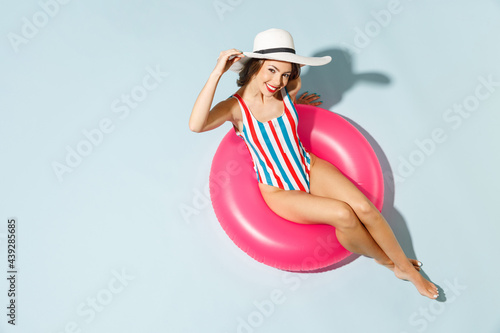 Top view young sexy woman slim body wear striped one-piece swimsuit widebrim hat lies inflatable tube ring hotel pool isolated on pastel blue color background Summer vacation sea rest sun tan concept photo