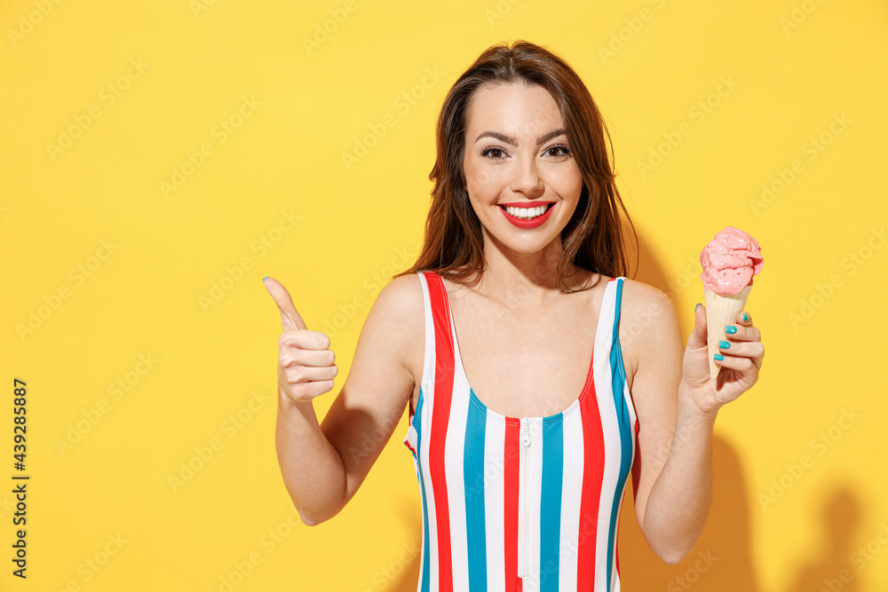 Happy young sexy woman slim body wear striped red blue one-piece swimsuit hold eat cold pink icecream isolated on vivid yellow color wall background studio. Summer hotel pool sea rest sun tan concept.