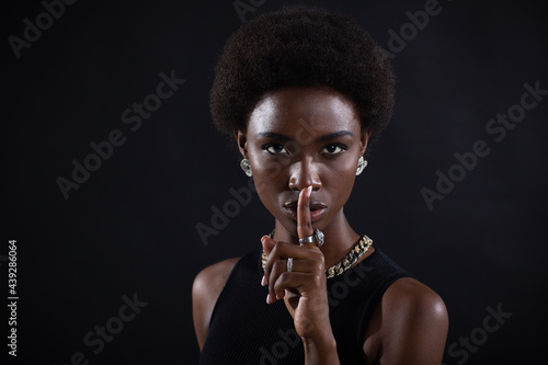 Closeup of beautiful young african american dark-skinned woman with finger on her lips showing shhh silence gesture on black background. photo