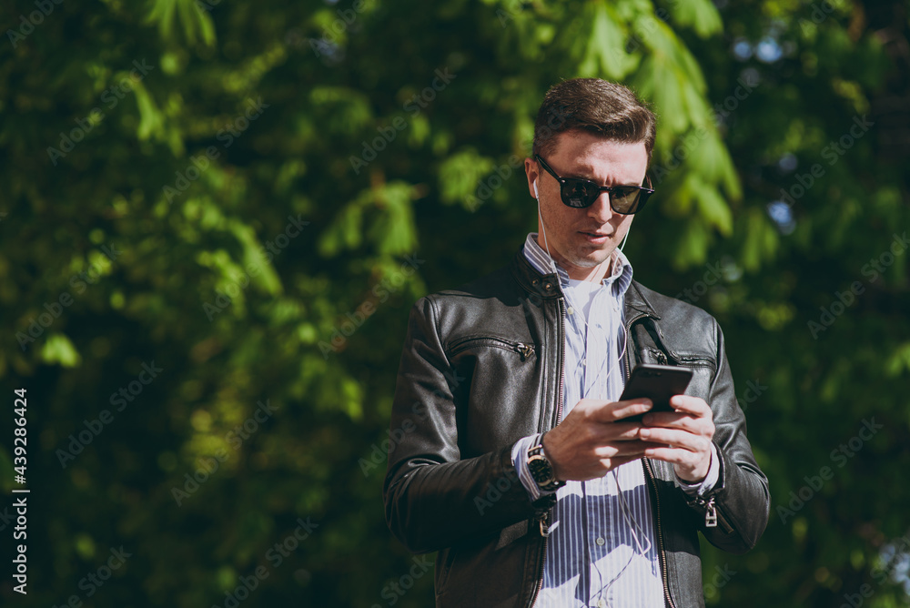 Fototapeta premium Young stylish caucasian man 20s wearing black leather jacket eyeglasses walk in green park in open air using mobile cell phone listen to music in earphones. Concept of urban lifestyle spring season