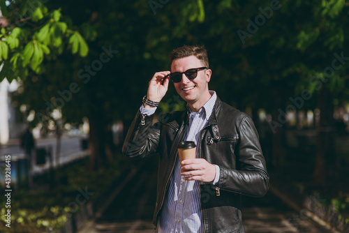 Young smiling happy man in black leather jacket eyeglasses stroll in green park hold takeaway delivery craft paper cup drink coffee to go, look camera. Concept of people urban lifestyle spring season.