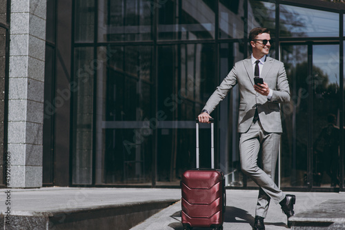Full length fun young employee business man 20s in grey suit stand near office glass wall building outdoors in downtown city hold mobile cell phone valise bag order booking taxi Business trip concept.