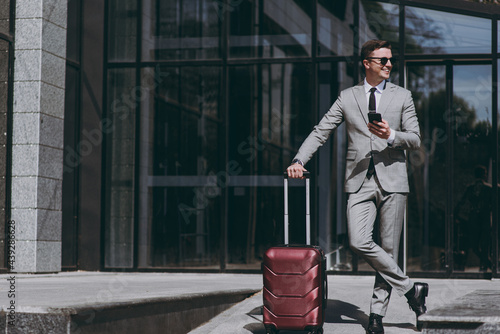 Cuadro en lienzo Full length fun young employee business man 20s in grey suit stand near office glass wall building outdoors in downtown city hold mobile cell phone valise bag order booking taxi Business trip concept