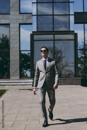 Full length young successful smiling employee business man 20s wearing grey suit walking go near office glass wall building outdoors in downtown city center look camera. Achievement career concept © ViDi Studio